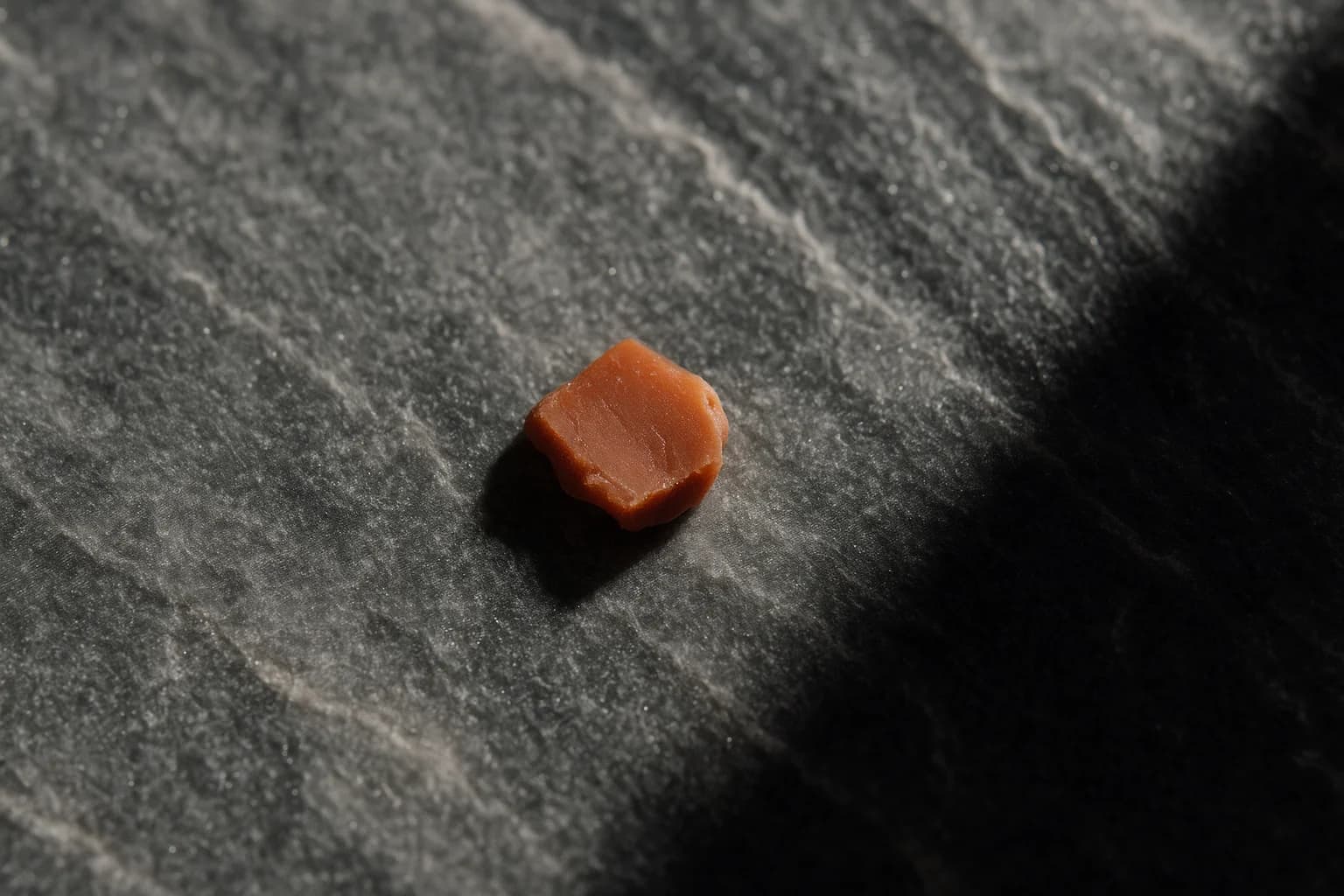 Extreme macro close-up of marble grey stone surface texture with a single small terracotta-colored wax seal fragment resting off-center in the lower third, evoking formal certification and document finality